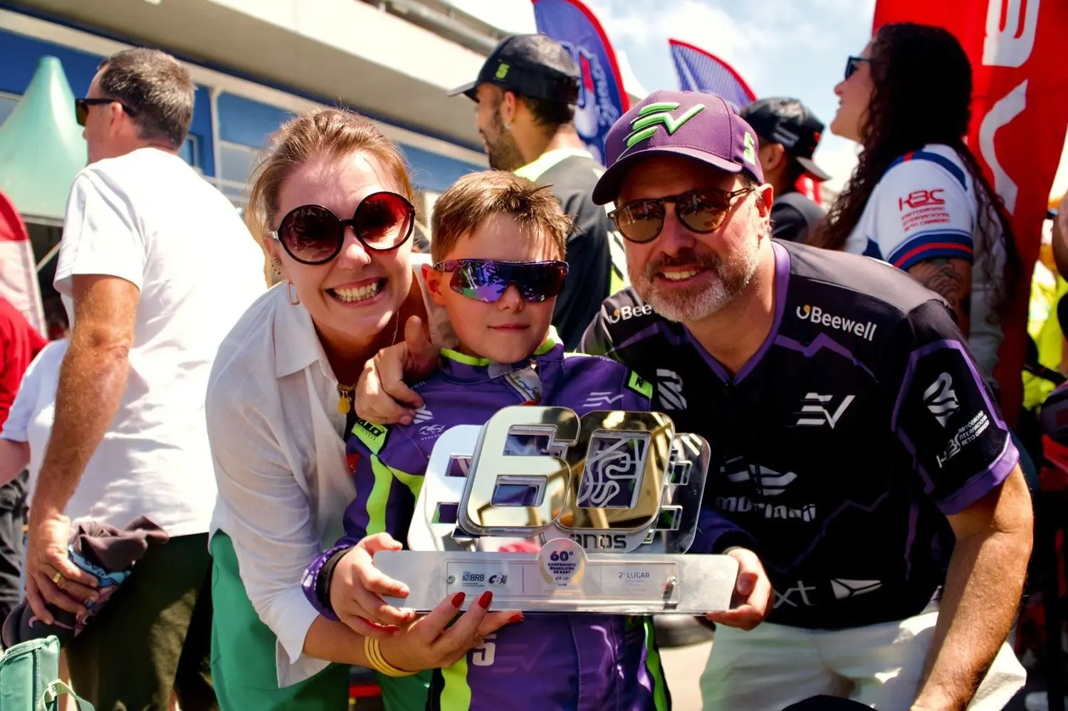 Enrico, de óculos escuros e macacão, segura um troféu ao lado de uma mulher e de um homem de boné, em um evento de kart