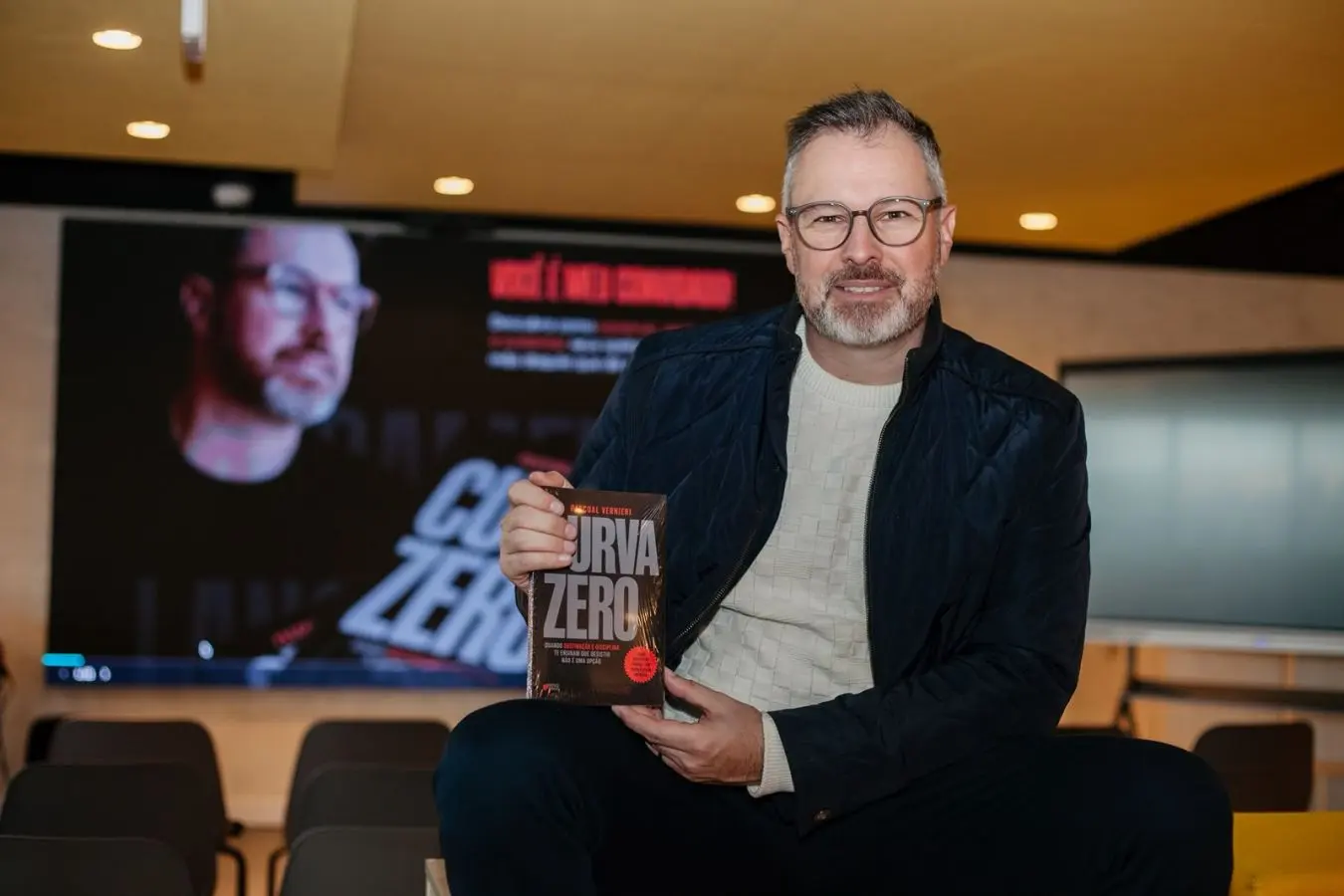 Pascoal Vernieri, de óculos, segurando o livro Curva Zero, com um telão ao fundo