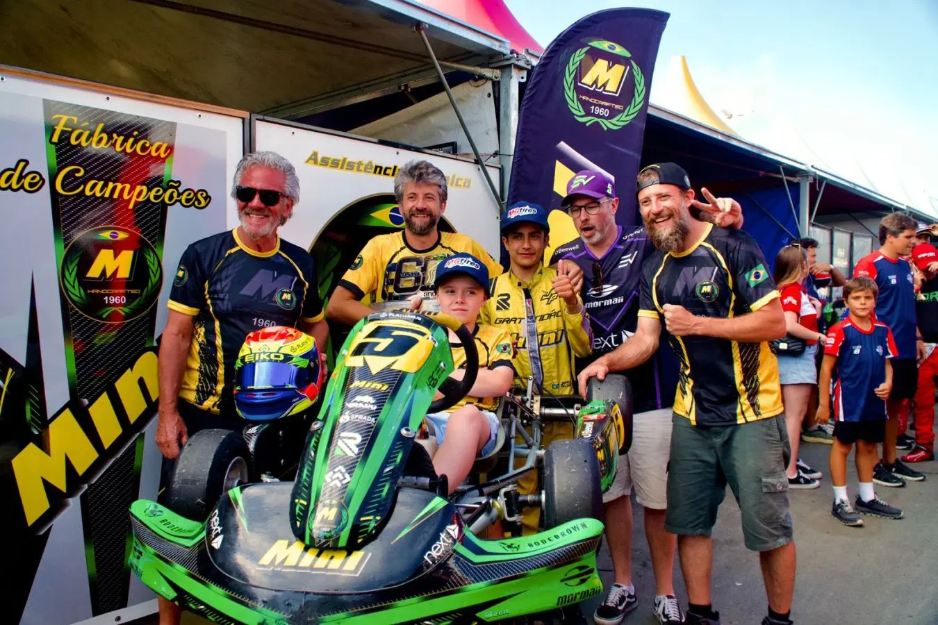 Enrico sentado em um kart verde, cercado por integrantes da equipe, nos boxes de uma prova