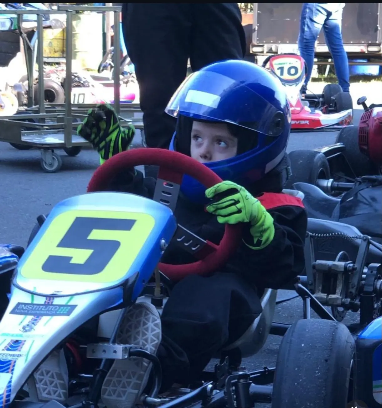 Enrico pequeno, de capacete azul e luvas verdes, ao volante de um kart com o número 5, nos boxes