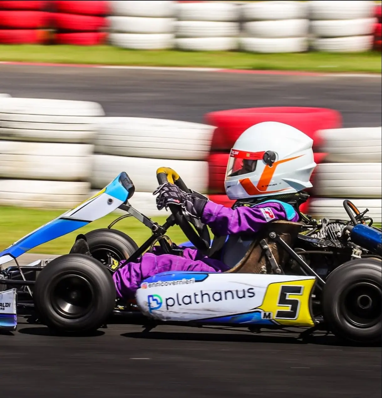 Enrico, de capacete branco e macacão roxo, pilotando um kart de lado em alta velocidade, com a pista e os pneus pintados desfocados ao fundo