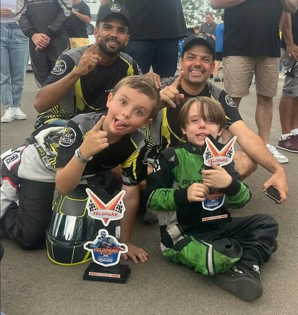Enrico, de macacão verde, sentado no chão com o troféu da Velopark Cup nas mãos, ao lado de outro menino e de dois homens de boné, todos sorrindo