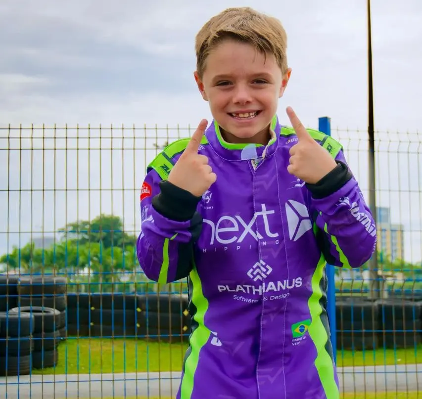 Enrico sorrindo, de macacão de corrida roxo com os logos Next e Plathanus no peito, apontando para cima com as duas mãos