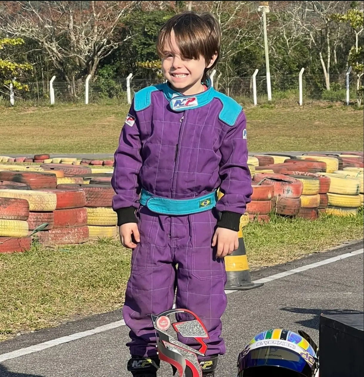Enrico ainda criança, de macacão de corrida roxo, sorrindo ao lado da pista, com capacetes aos pés